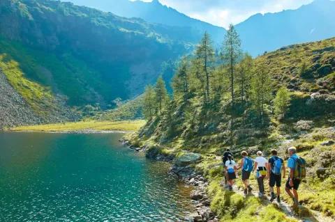 Eine Gruppe von fünf Wanderern geht auf einem schmalen Pfad direkt entlang eines blauen Bergsees. Hinter dem See ist ein hügeliges Tal zu sehen.