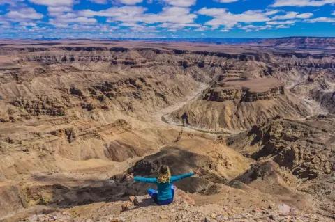 Eine Frau sitzt im Schneidersitz am Rand eines weiten Canyons. Sie steckt die Arme seitlich aus und blickt über den Canyon.