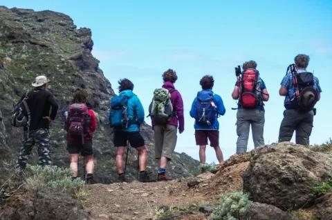 Sieben Wanderer stehen mit einem Tagesrucksack auf einer Anhöhe und blicken in Richtung Horizont. Die Wanderer stehen nebeneinander und mit dem Rücken zur Kamera