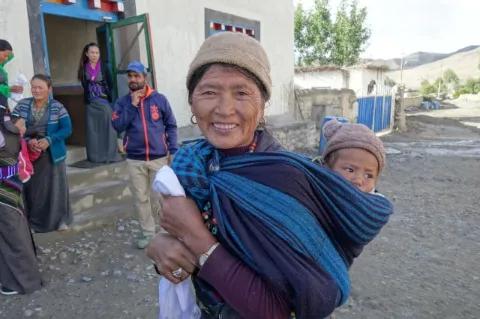 Eine nepalesische Frau hat ein blaues Tragetuch mit einem Baby auf dem Rücken und grinst in die Kamera.