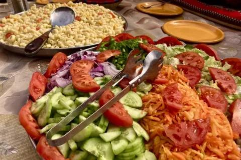 Auf einem Tisch steht eine große Schüssel, die mit geschnittenen Tomaten, Karotten und Gurken gefüllt ist. Im Hintergrund ist eine weitere Schüssel mit Nudeln zu sehen.