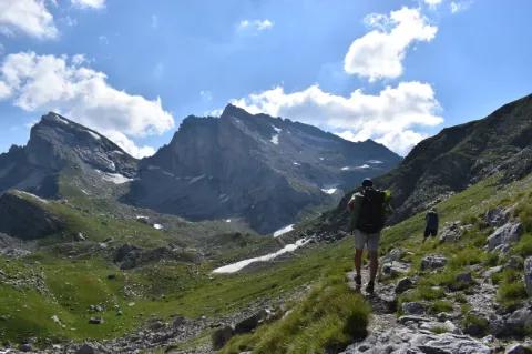 Ein Wanderer geht auf einem schmalen Pfad durch hügelige Landschaften. Einige Altschneefelder sind zu sehen.