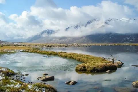 Ein hellblauer Fluss fließt auf einer flachen Ebene durch eine grüne Ebene. Im Hintergrund ist eine hügelige Landschaft zu sehen. Am Himmel sind Wolken zu sehen.