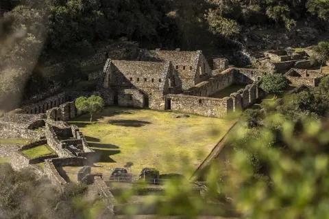 Blick auf die Ruine einer alten Inkastätte aus Steinen. Es sind noch zwei Gebäude mit einem Steindach und mehrere Mauerreste zu sehen. Die Stätte ist von dichtem Wald umgeben.
