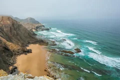 Weiter Blick auf eine Küstenlandschaft. Das türkise Meer schlägt an einen Sandstrand hinter dem eine steile Felswand steht.