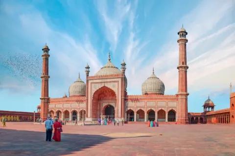Es ist die Frontalansicht einer großen Moschee mit zwei hohen Türmen und runden Dächern zu sehen. Die verwendeten Steine sind rot. Vor der Moschee stehen zwei Menschen.