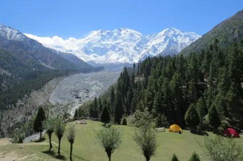 Im Vordergrund ist eine grüne Wiese mit zwei Zelten zu sehen. Hinter einer felsigen Tallandschaft ist das schneebedeckte Bergmassiv des Nanga Parbat zu sehen.