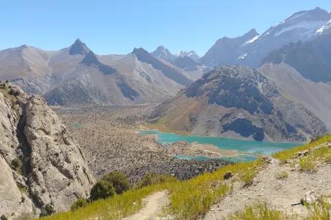 Aus der Distanz ist ein hellblauer, kleiner See zu sehen, der an eine felsige, karge Ebene angrenzt. Im Hintergrund sind spitz zulaufende Hügel zu sehen, die an manchen Stellen mit kleinen Waldstücken bewachsen sind.