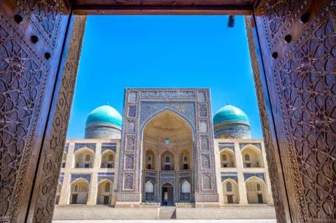 Blick durch ein massives, mit Ornamenten verziertes Tor auf eine Moschee mit blauem Mosaik. Links und rechts neben dem hohen Eingang sind zwei blaue Kuppeldächer zu sehen.