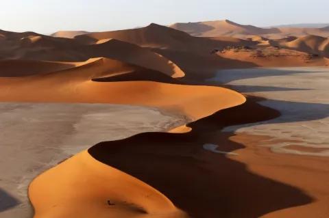 Blick auf eine geschwungene, steil zulaufende Düne, die in Kurven verläuft. Auf der rechten Seite der Düne wirft diese einen großen Schatten. Im Hintergrund sind weitere Sanddünen zu sehen.