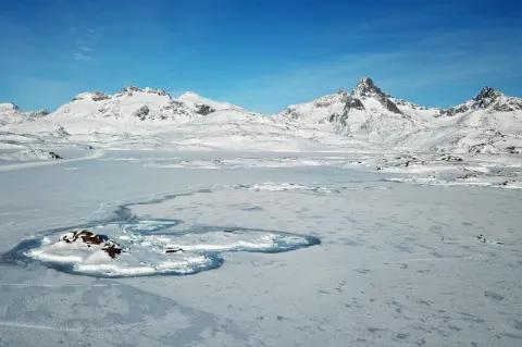 Blick über eine flache, große Eisfläche die mit Schnee bedeckt ist. Auf der Fläche ist eine kleine Siedlung zu sehen. Im Hintergrund erheben sich schneebedeckte, spitze Felsen.