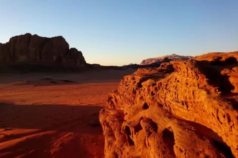 Blick über eine rote Sandstein und Sandlandschaft, die von der untergehenden Sonne angestrahlt wird und deshalb in goldgelbem Licht erstrahlt.