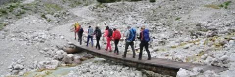 Eine Gruppe von acht Wanderern geht über eine kleine Holzbrücke. Der Untergrund ist felsig.