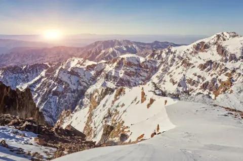 Blick über die Gebirgslandschaft im Winter. Die Sonne geht gerade am Horizont unter.