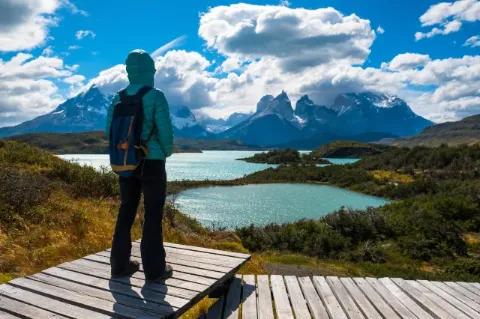 Eine Frau mit Rucksack und Kapuze steht mit dem Rücken zur Kamera auf einem Holzsteg. Sie blickt auf zwei hellblaue Seen, die in einer grünen Landschaft liegen. Im Hintergrund sind die Berge des Torres del Paine Nationalpark zu sehen. Der Himmel ist blau und von Wolken durchzogen.