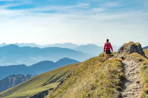 Eine Wanderin mit roter Jacke wandert auf einem steinigen Pfad auf einem Grat. Im Hintergrund ist schemenhaft ein Gebirge zu sehen.