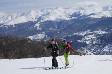 Zwei Skitourengeher gehen auf einer weißen Schneedecke und blicken in die Kamera. Im Hintergrund ist ein verschneites Gebirge zu sehen.