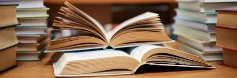 Zwei aufgeschlagene Bücher liegen auf einem Tisch und sind von weiteren Bücherstapeln umgeben