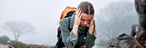 Eine junge Frau sitzt auf einem Felsen und hält sich mit beiden Händen links und rechts den Kopf.