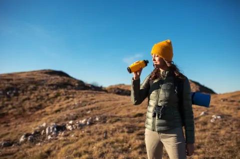 Eine junge Frau mit gelber Mütze hält eine gelbe Wasserflasche in der Hand und hat die Augen geschlossen.