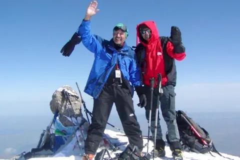 Zwei Wanderer stehen freudestrahlend auf einem schneebedeckten Gipfel. Ihre Ausrüstung liegt vor ihnen.