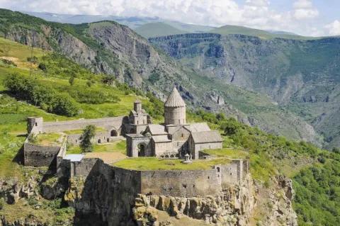Ein Kloster im Hochgebirge. Die Berglandschaft ist felsig und grün. Das Kloster ist aus hellbraunem Stein.