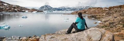Eine Wanderin sitzt auf einem Felsen und blickt aufs Wasser. In der Ferne sind schneebedeckte Berge.