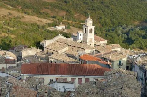 Ein Dorf an einem Hang. Die Gebäude sind aus hellbraunem Stein. In der Mitte ist eine Kirche.