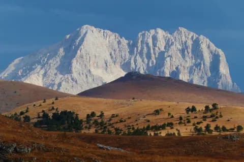 Ein großer, grau-weißer Berg am Horizont. Davor sind kleinere orange-braune Berge mit Bäumen.