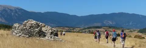 Wanderer gehen einen Pfad entlang. Links ist ein Steinhügel, am Horizont ein Bergpanorama.