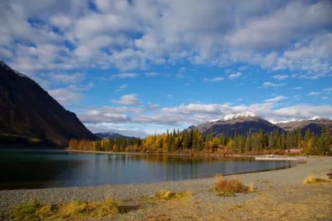 Ein See mit einem trockenen Ufer und Bergen. Die Blätter der Bäume im Wald färben sich orange.