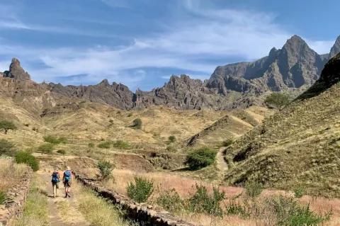 Zwei Wanderer laufen im Tal einen Pfad entlang. Im Hintergrund sind Berge.
