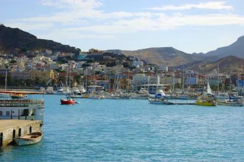 Mehrere Boote sind auf dem Wasser am Hafen. Dahinter ist eine Stadt und eine Berglandschaft.
