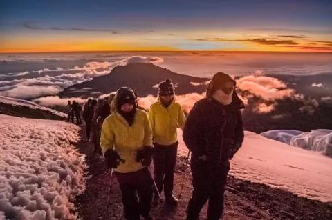 Mehrere dick eingepackte Wanderer steigen auf einen Berg hinauf. Die Sonne geht gerade auf.