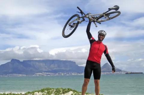 Ein Radfahrer steht an der Küste vor den türkisen Meer und hält sein Fahrrad über seinen Kopf.