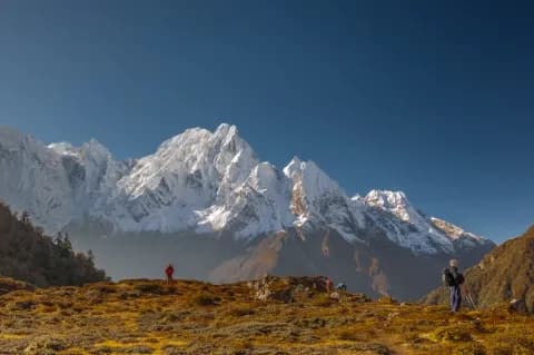 Zwei Wanderer blicken von einem orange-grün bewachsenen Berg auf einen schneebedeckten Berg.