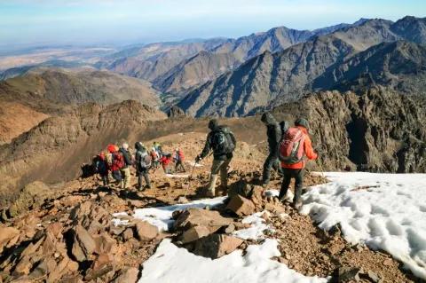 Mehrere Wanderer steigen einen Berg herab. Die Landschaft ist felsig und teils schneebedeckt.