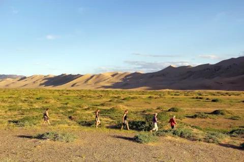 Fünf Wanderer gehen durch ein grün bewachsenes Tal. Im Hintergrund sind die braunen Berge.