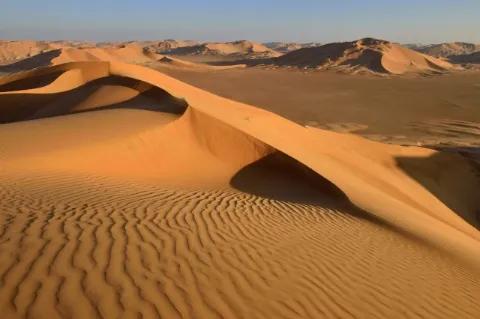 Eine Wüstenlandschaft mit rot-orangenen Dünen. Der Himmel ist blau.
