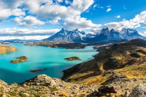 Ein blauer See umgeben von Hügeln und hohen, schneebedeckten Bergen. Am Himmel sind Wolken.