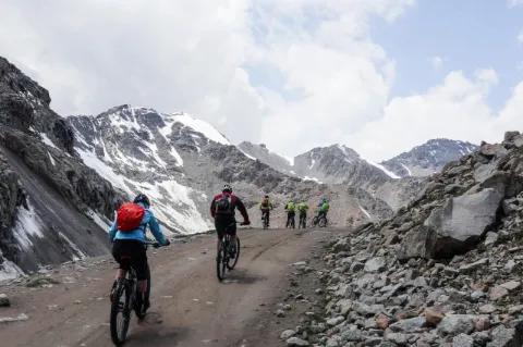 Mehrere Radfahrer fahren im felsigen, teils schneebedeckten Gebirge. Am Himmel sind Wolken.