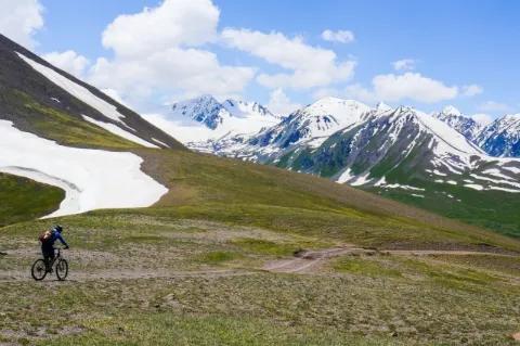Ein Fahrradfahrer fährt durch ein grünes Tal. Hinter ihm sind teils schneebedeckte Berge.