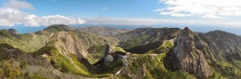 Eine grün bewachsene Berglandschaft. In der Ferne ist das Meer. Am Himmel sind einzelne Wolken.