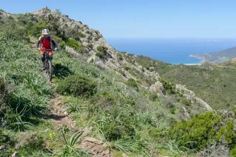 Ein Radfahrer fährt über einen schmalen Pfad auf einem Berg in Sardinien. Im Hintergrund ist das Meer zu sehen.