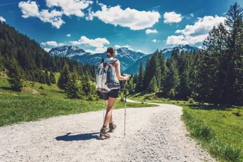 Eine Frau mit Stöcken steht auf einem Kiesweg der von Bäumen umgeben ist und in Richtung Berge führt.