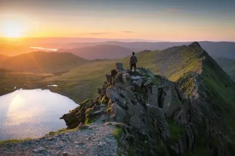 Ein Wanderer steht auf einem schmalen Grad und schaut in Richtung Sonnenaufgang. Links neben dem Grad ist ein Bergsee zu sehen.