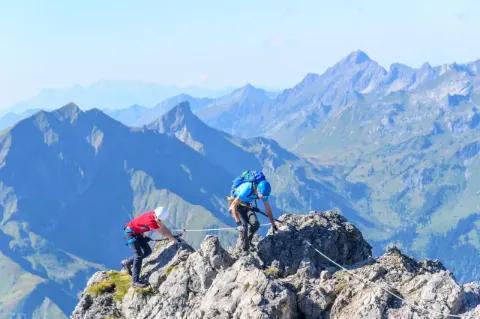 Zwei Menschen mit Kletterausrüstung klettern entlang eines Stahlseils auf felsigem Untergrund.