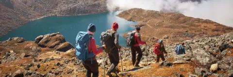 Fünf Wanderer steigen einen Berg ab. Der Boden ist rötlich und geröllig. Im Tal ist ein Bergsee.