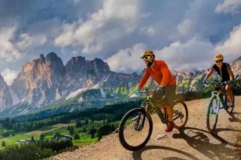 Ein Paar radelt auf E-Bikes auf einer Schotterpiste vor grünen Wiesen und einer Bergkulisse im Hintergrund bei bewölktem Himmel