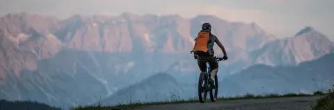 Mountainbiker mit Rucksack fährt über eine Teerstraße mit Blick auf die Bergkette bei leicht trübem Wetter.